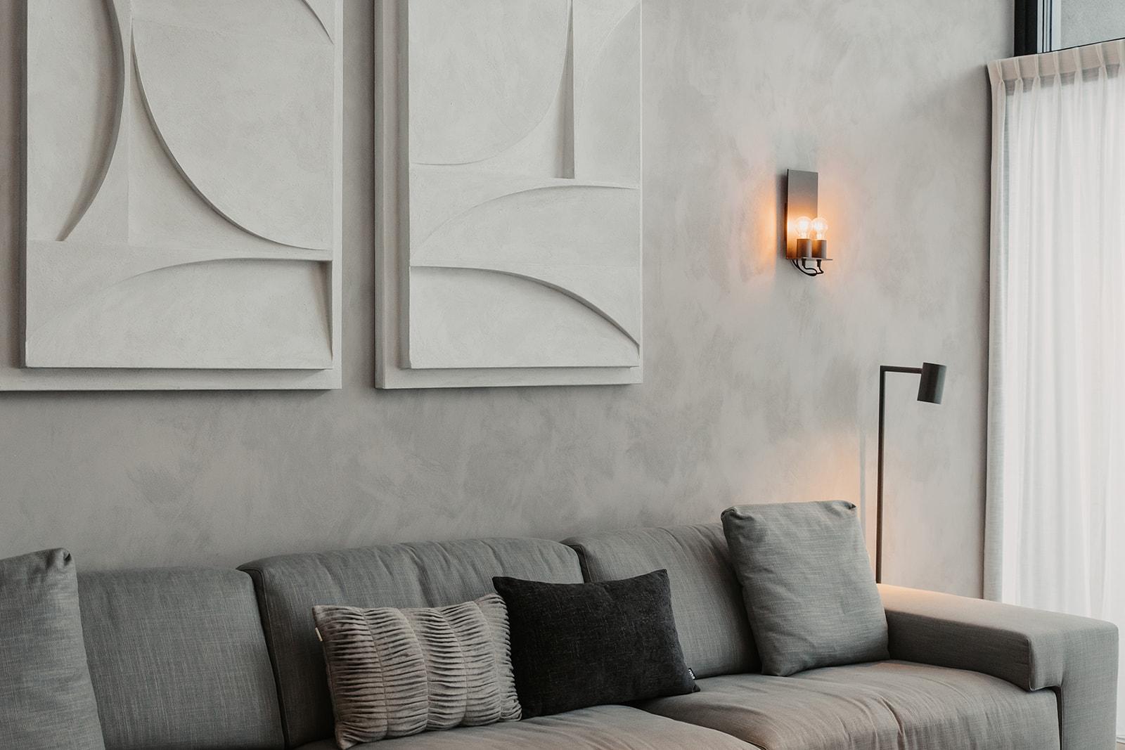 A modern living room with a gray sofa and several cushions. Two abstract artworks hang on the wall and there is an elegant light source.