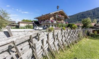 A beautiful Alpine house with blooming balconies. In the foreground, there is a wooden fence, and in the background, mountains can be seen.