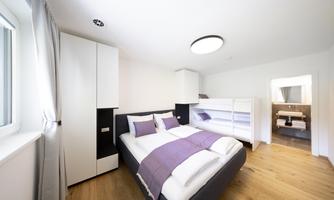 A modern bedroom with a double bed and a bunk bed. The room has bright walls, large windows, and a minimalist style.