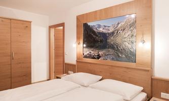 A modern bedroom with a large wooden wardrobe and a double bed. On the wall hangs an impressive picture of a mountain landscape with a clear lake.