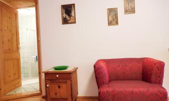 A cozy living room with a red armchair and a small wooden sideboard. In the background, you can see an open door to the bathroom.