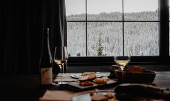 A cozy table with food and wine glasses, the view shows snowy trees through a window. The atmosphere is warm and inviting.