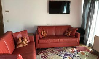 A cozy living room with red sofas and decorative cushions. A TV hangs on the wall, and the room has a colorful carpet.