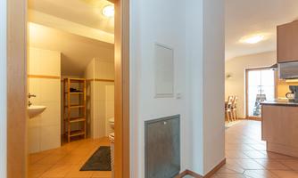 A spacious hallway with an open bathroom door and bright tiles. On the left, shelves are visible and on the right, access to a living room with a kitchen area.