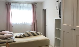 A cozy bedroom with a large bed and colorful pillows. The room has bright walls, a carpet, and a window with curtains.