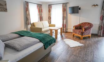 A modern room with a bed, a sofa, and an armchair. Large windows let in plenty of daylight.
