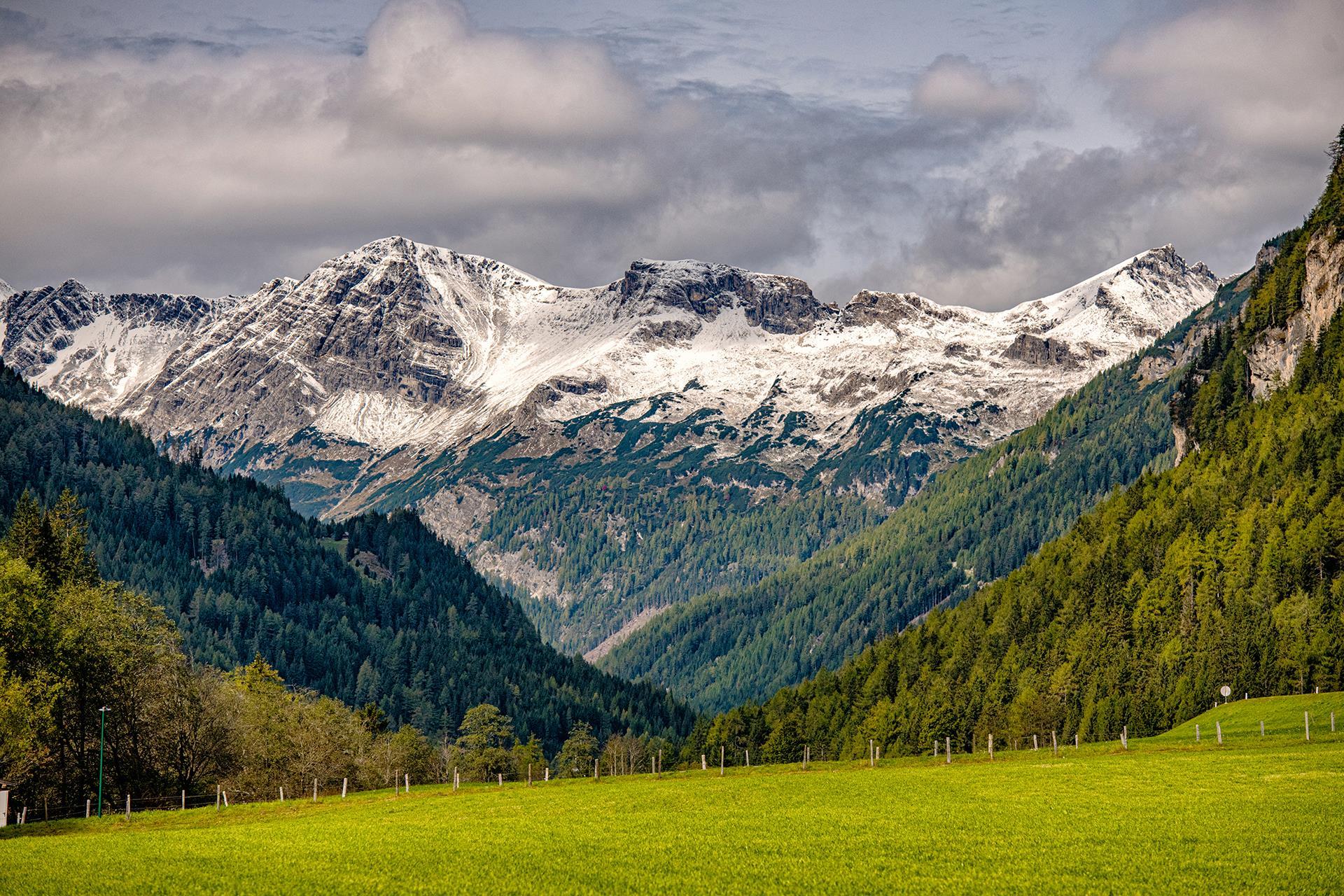 An impressive mountain landscape with snow-covered peaks and green forests in the foreground. The sky is partly cloudy, adding a dramatic touch to the scene.