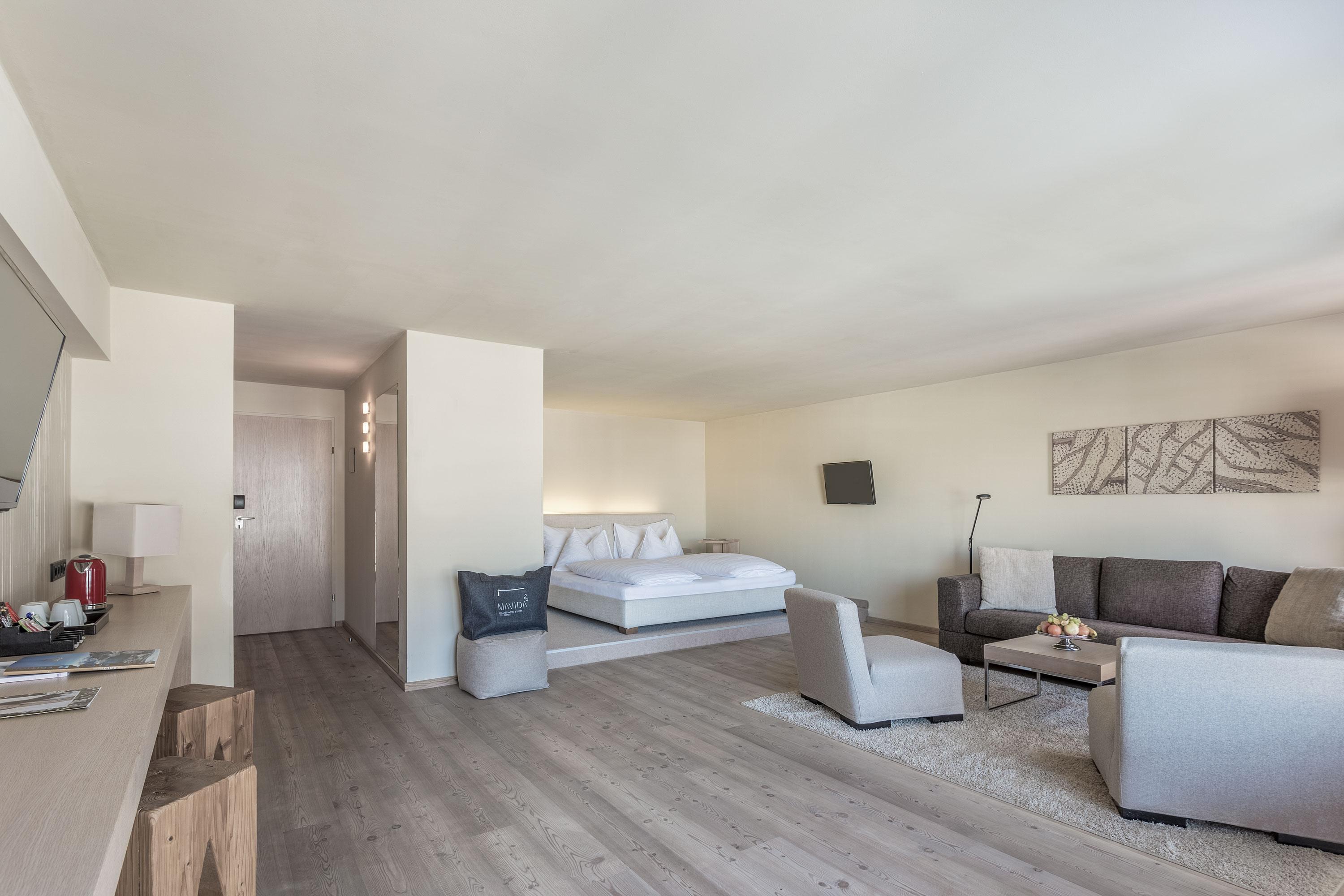 A modern hotel room with a cozy living area and a large bed. Bright walls and wooden flooring create an inviting atmosphere.