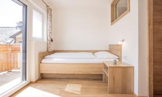 A bright bedroom with a wooden bed and a bedside table. Large windows offer a view of a balcony.