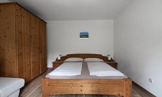 A simple bedroom with a wooden bed and two pillows. The room contains a large wardrobe and a small picture on the wall.