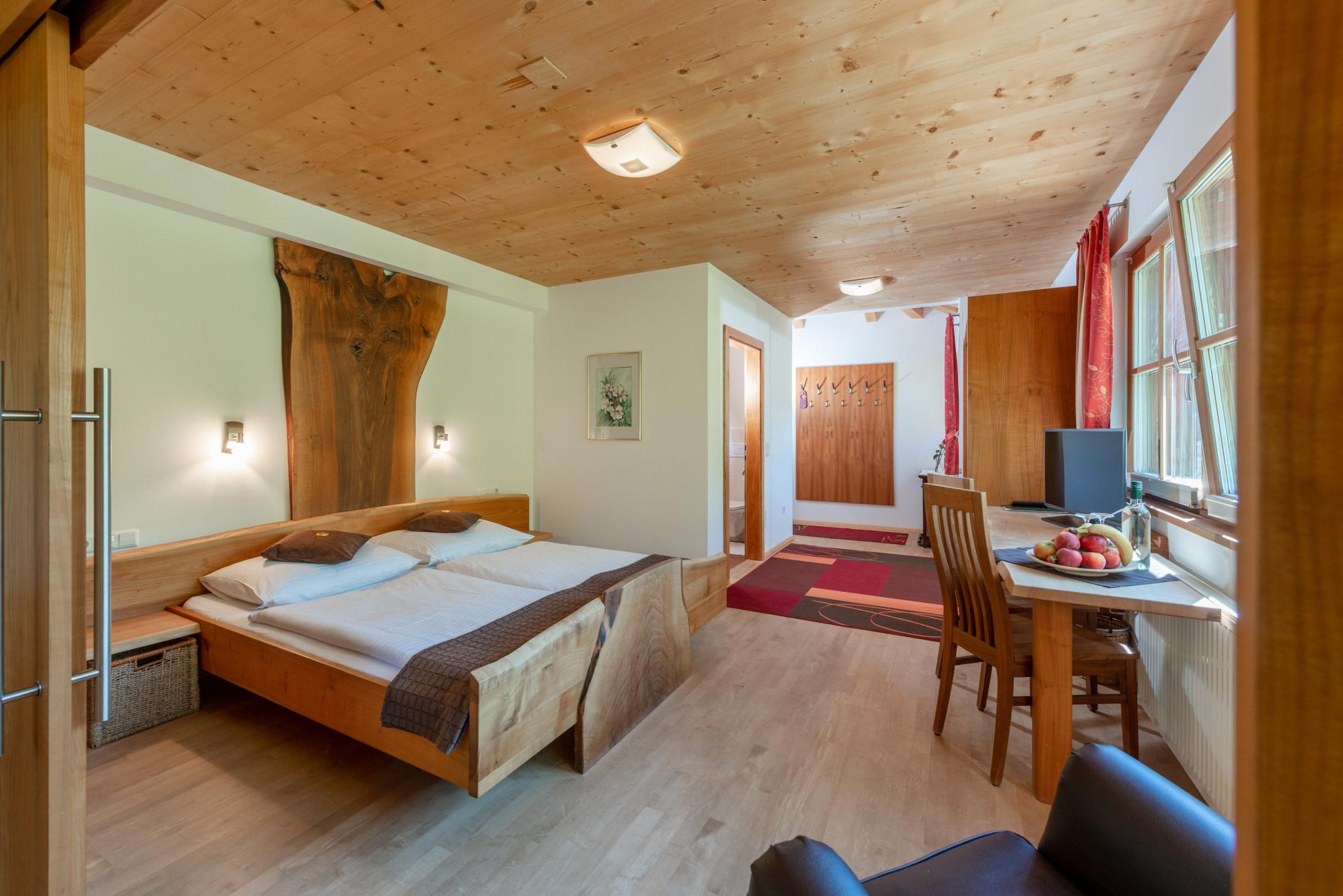 A cozy room with a double bed and wooden decor. There is a desk with fruit and a chair, as well as large windows for plenty of daylight.