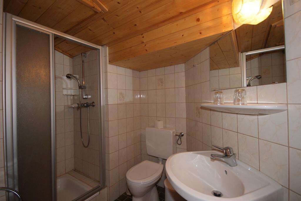 A small bathroom with a shower, sink, and toilet. The walls are tiled, and the ceiling is made of wood.