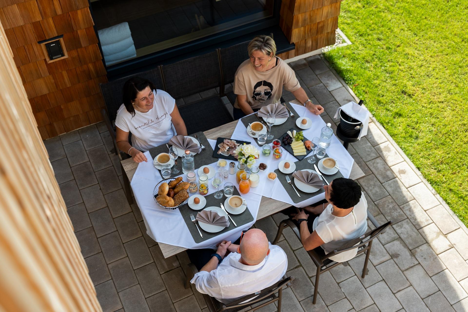 A group of four people is sitting at an outdoor table enjoying breakfast. The table is set with various foods and drinks while the sun is shining.