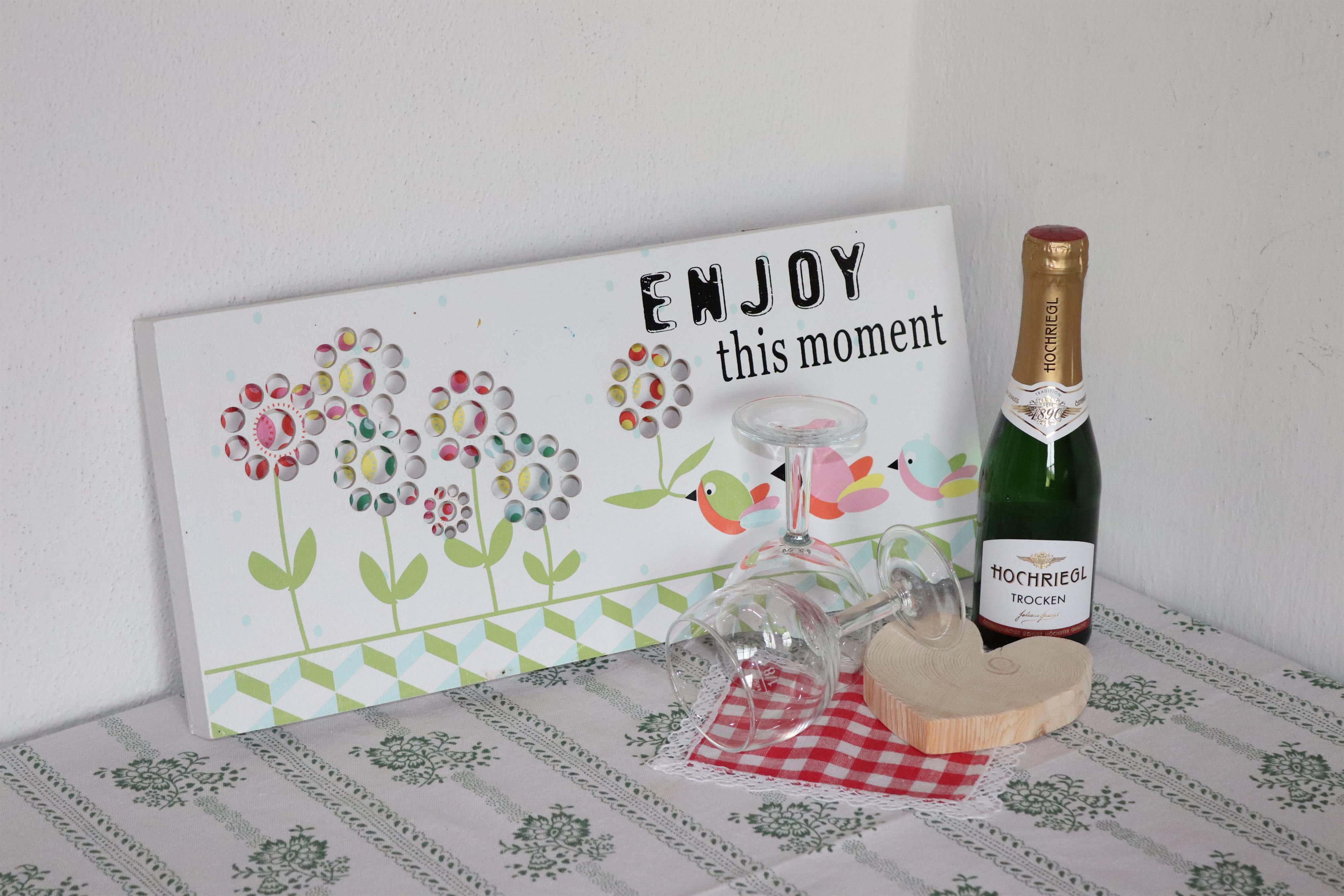 A decorative sign reading "ENJOY this moment" with colorful flower motifs. Next to it are a bottle of champagne, a glass, and a piece of cheese on a table with a checkered cloth.