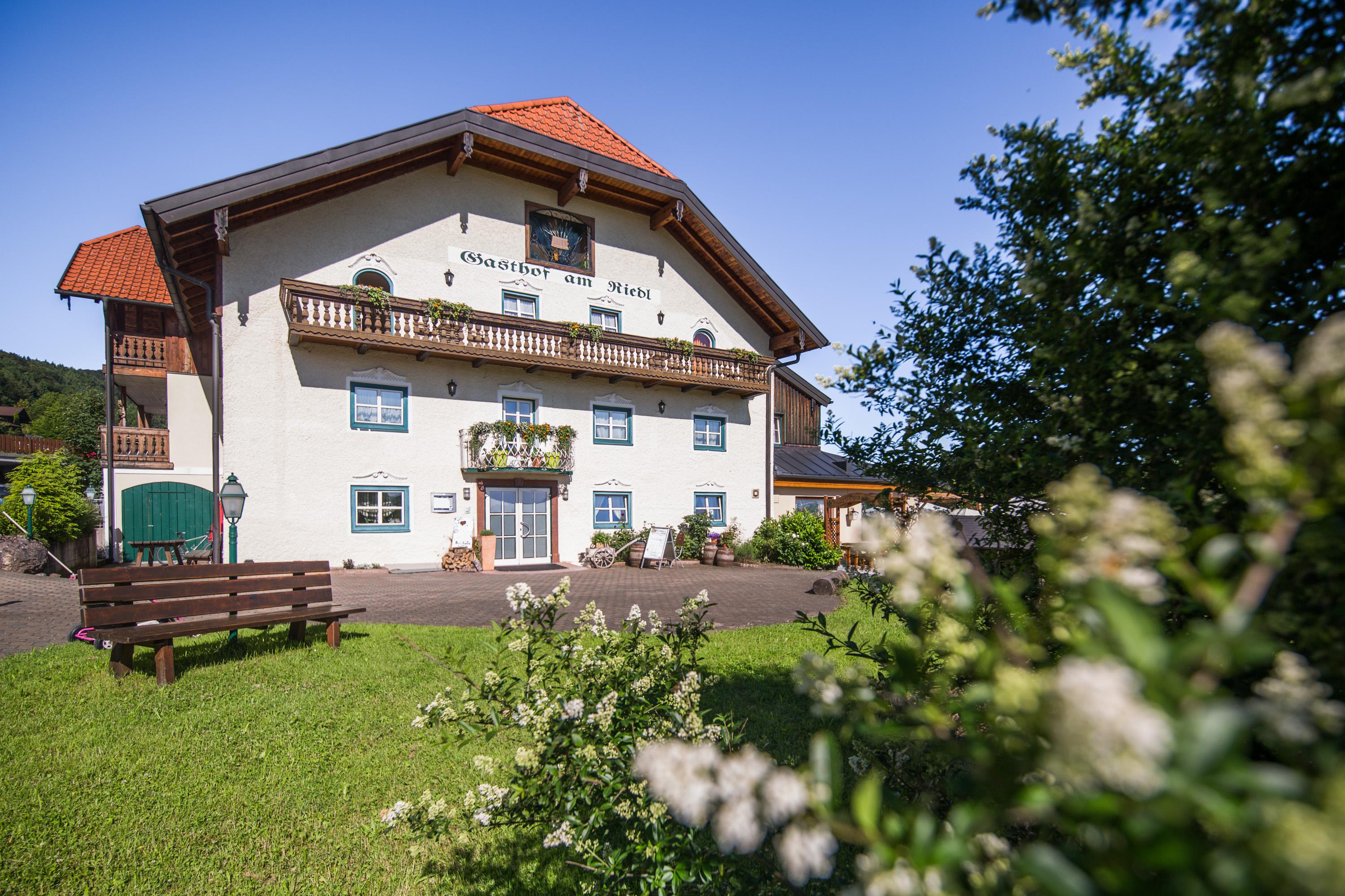 A charming building with a red roof and an inviting entrance area. The garden is green and well-kept, surrounded by trees and bushes.