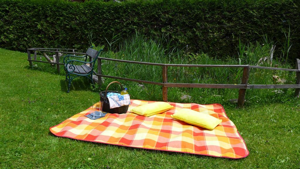 A cozy picnic blanket lies on the green grass. Next to the blanket is a basket with snacks and two yellow pillows.