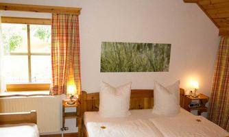 A cozy bedroom with two beds, a small window, and warm wooden interiors. The walls are decorated with simple curtains, and a picture of grasses hangs on the wall.