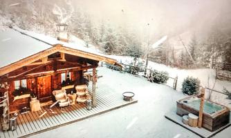 A cozy wooden house in a snowy landscape. In the foreground, a terrace and a hot tub can be seen.