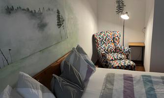 A cozy bedroom with a large bed and a colorful armchair. On the wall hangs a piece of artwork featuring tree motifs.