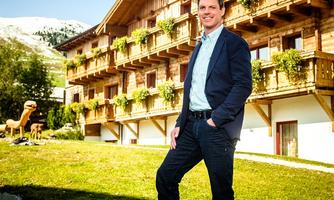 A man stands smiling in front of a traditional wooden building in a mountainous setting. The architecture is rustic, with balconies and green plants.