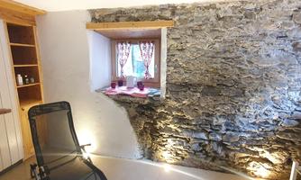 A cozy room with a stone wall and a window dressed with curtains. There is a comfortable chair and some decorative elements on the windowsill.