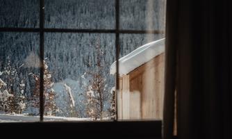 A view from a window onto a snowy landscape with a wooden house. The trees are covered in snow and the mountains in the background are visible.