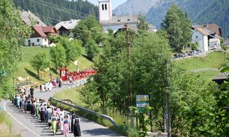 A festive parade with people in traditional clothing passes through a picturesque landscape. In the background, there are houses and mountains.