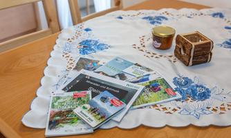 On a table lie several brochures next to a small jar of honey and a decorative wooden box. The tablecloth has a blue floral pattern.