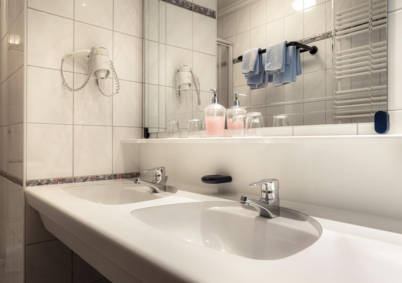 A modern bathroom with two sinks and large mirrors. The walls are tiled and towels as well as soap dispensers are visible.