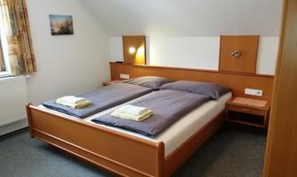 A simple bedroom with two single beds, bedding in blue, and a small bedside lamp. The walls are light, and there is a window with curtains.