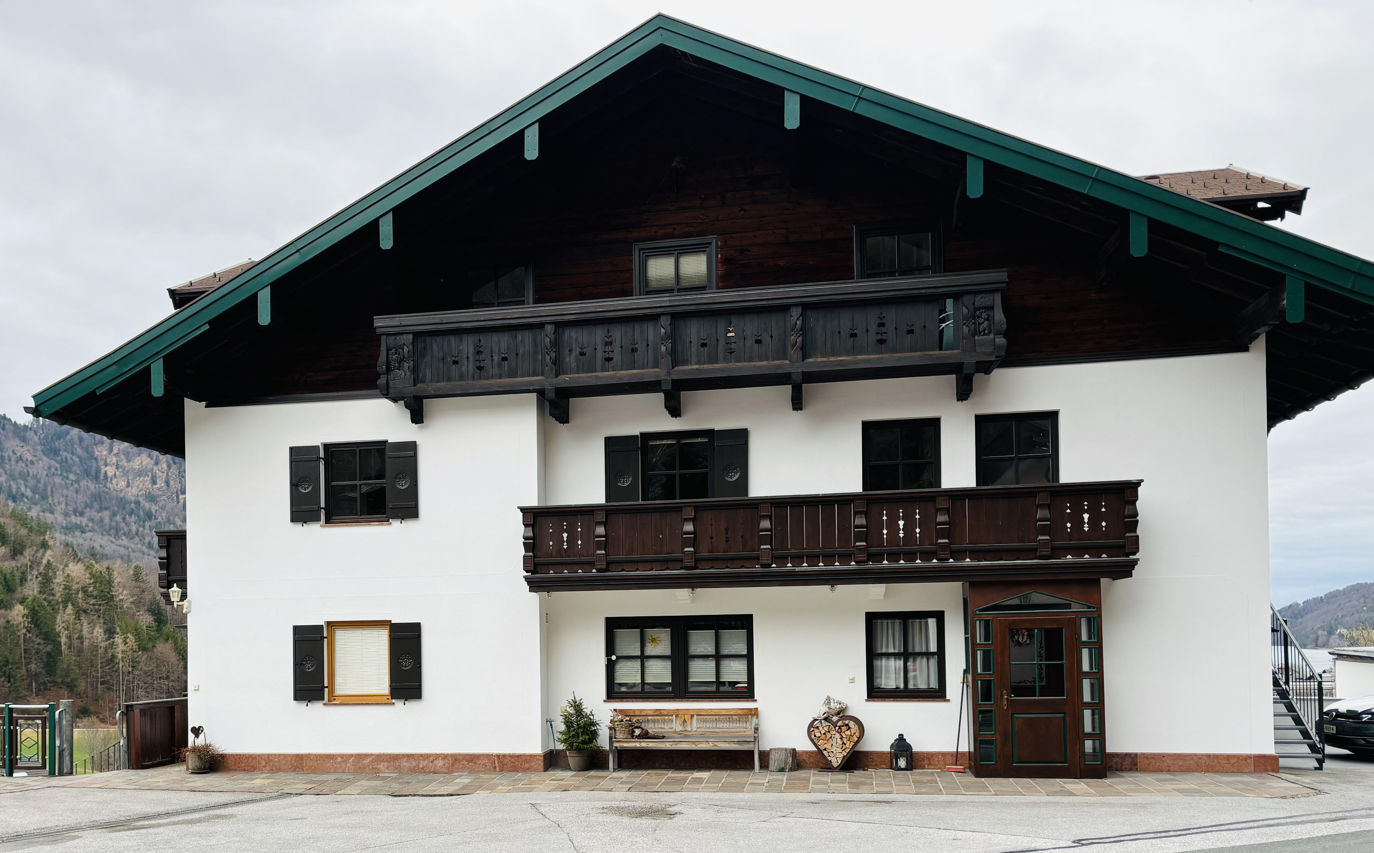 A traditional two-story country house with a wooden balcony and a green roof. It is located in a rural setting, surrounded by mountains.