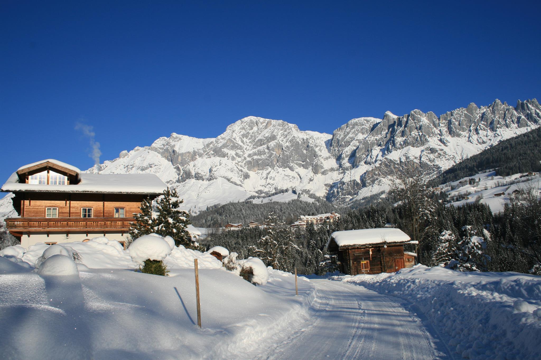 A picturesque winter village with snow-covered hills and majestic mountains in the background. The clear blue skies give the scene a calm atmosphere.