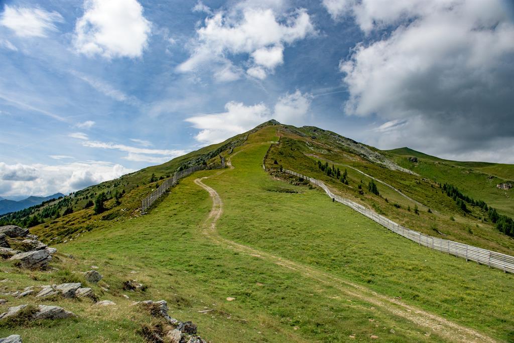 A green mountain landscape with a clear path and wooded slopes. The sky is partly cloudy and radiates a peaceful atmosphere.