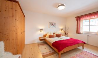A cozy bedroom with a large double bed and warm wood interiors. Large windows with bright curtains let in plenty of light.