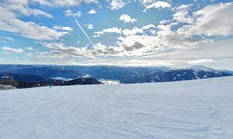 A snowy landscape with gentle hills and impressive mountains in the background. The sky is blue with a few clouds and rays of sunshine.