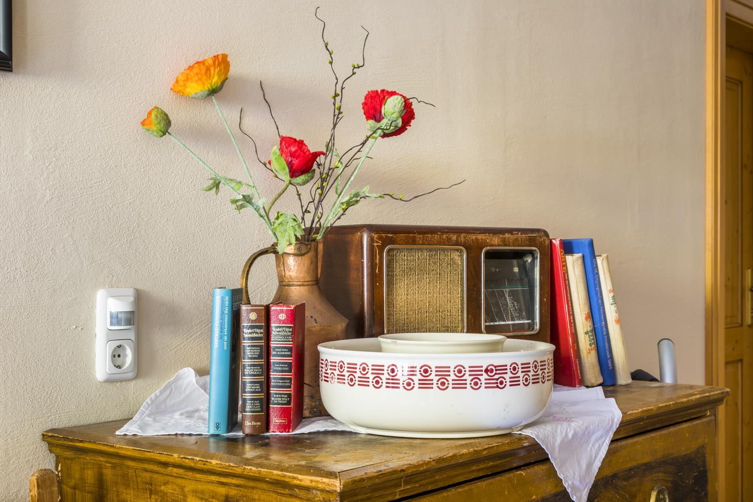 A wooden cabinet with a vintage radio, a porcelain bowl, and colorful flowers in a vase. Next to the radio are several books in different colors.