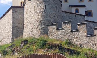 An impressive castle with a stone tower and historic masonry. In the foreground, there is a modern wooden sculpture that is harmoniously integrated into the surroundings.