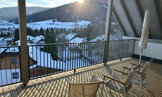 A beautiful terrace with a view of snow-covered mountains and a sunny sky. Two chairs and a sunshade are ready for relaxed hours outdoors.
