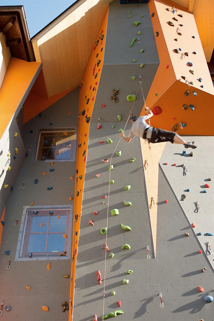 A climbing wall with many colorful grips and a climber floating high above. The wall is orange and gray and is located on a building.