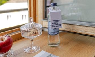 A glass bottle of water stands next to a transparent dessert bowl on a wooden table. A red apple and a magazine are also lying on the table.