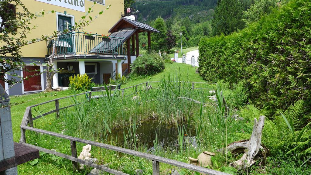 A quiet garden with a small pond and green plants. In the background, there is a building with a balcony.