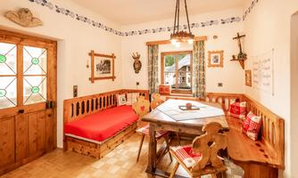 A cozy dining area in Alpine style with a large table and a bench. The walls are adorned with traditional decorations and windows featuring colorful glass.
