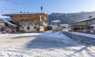 A scenic alpine landscape with snow-covered houses and mountains in the background. The sky is clear and blue, creating a tranquil winter atmosphere.