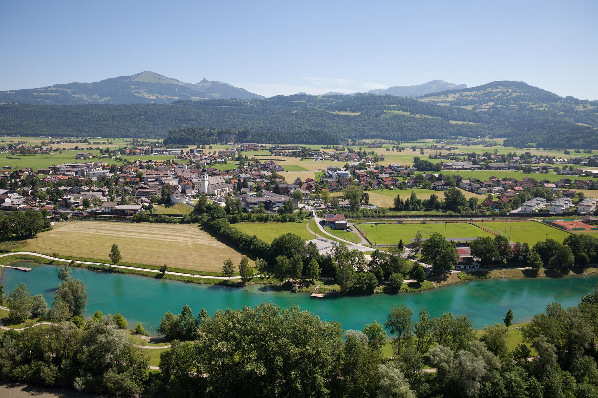 A picturesque landscape with a calm river and green meadows. In the background, gentle hills and mountains can be seen.