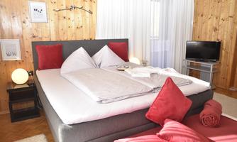 A cozy bedroom with a large bed and red pillows. Natural light shines through the curtains, and a TV is in the background.