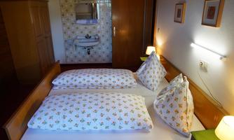 A cozy bedroom with two single beds and decorative pillows. In the background, a sink and some wall pictures are visible.