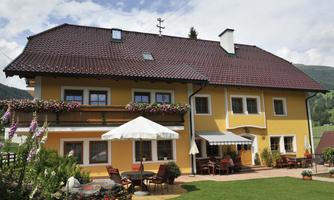 A cozy, yellow house with a red roof and blooming windows. In the foreground, there is a green lawn area and seating under a sun umbrella.