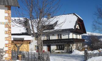 A traditional, snow-covered house with a balcony and wooden facade. In the foreground are a fence and bare trees under a clear sky.