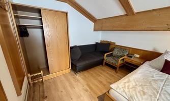 A cozy room with wood paneling and a large window. There is a bed, a couch, and a wardrobe.