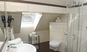 A modern bathroom with a shower and a sink. The walls are light tiled and a large window provides natural light.